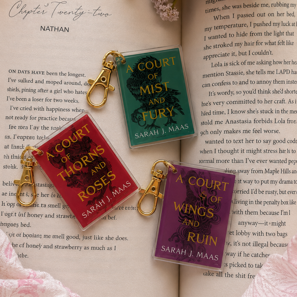 Three colorful book-themed keychains with gold clasps resting on an open book surrounded by pink roses. The keychains feature red, green, and purple covers with gold and white text.