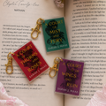Three colorful book-themed keychains with gold clasps resting on an open book surrounded by pink roses. The keychains feature red, green, and purple covers with gold and white text.